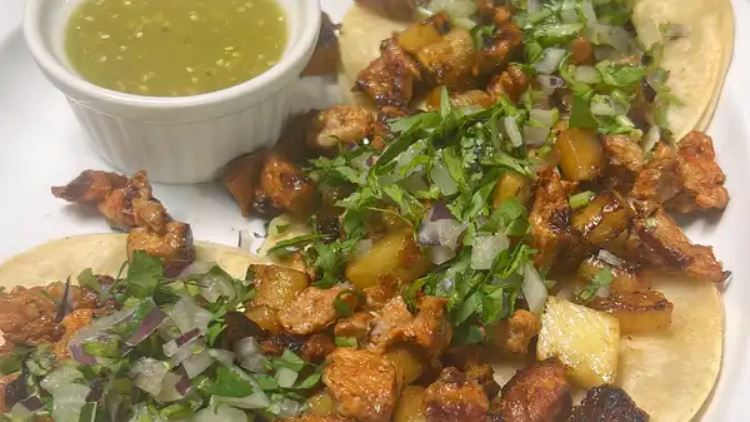 Two open faced al pastor tacos with grilled pineapple, cilantro and onion, next to a ramekin of green tomatillo salsa