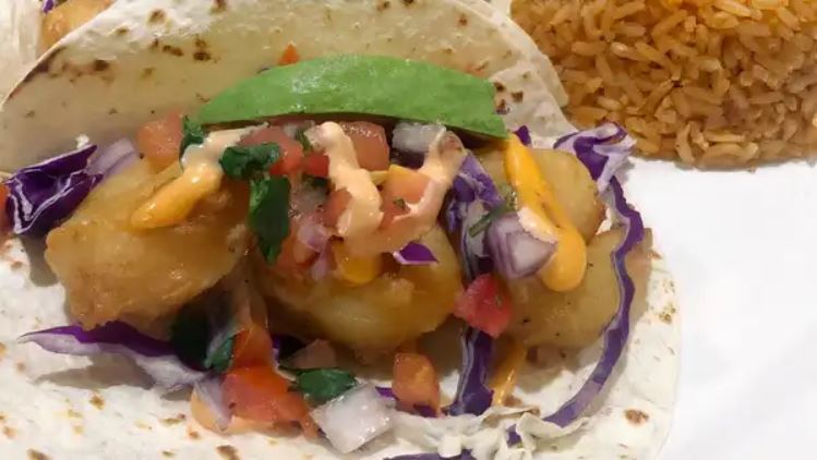 A baja shrimp taco on a flour tortilla with red cabbage, pico, avocado and a drizzle of bang bang sauce, beside Mexican rice