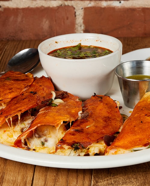 Four birria quesa tacos with cheese pulling at the edges, a white bowl of consome and a cup of salsa, on a wooden table against exposed red brick