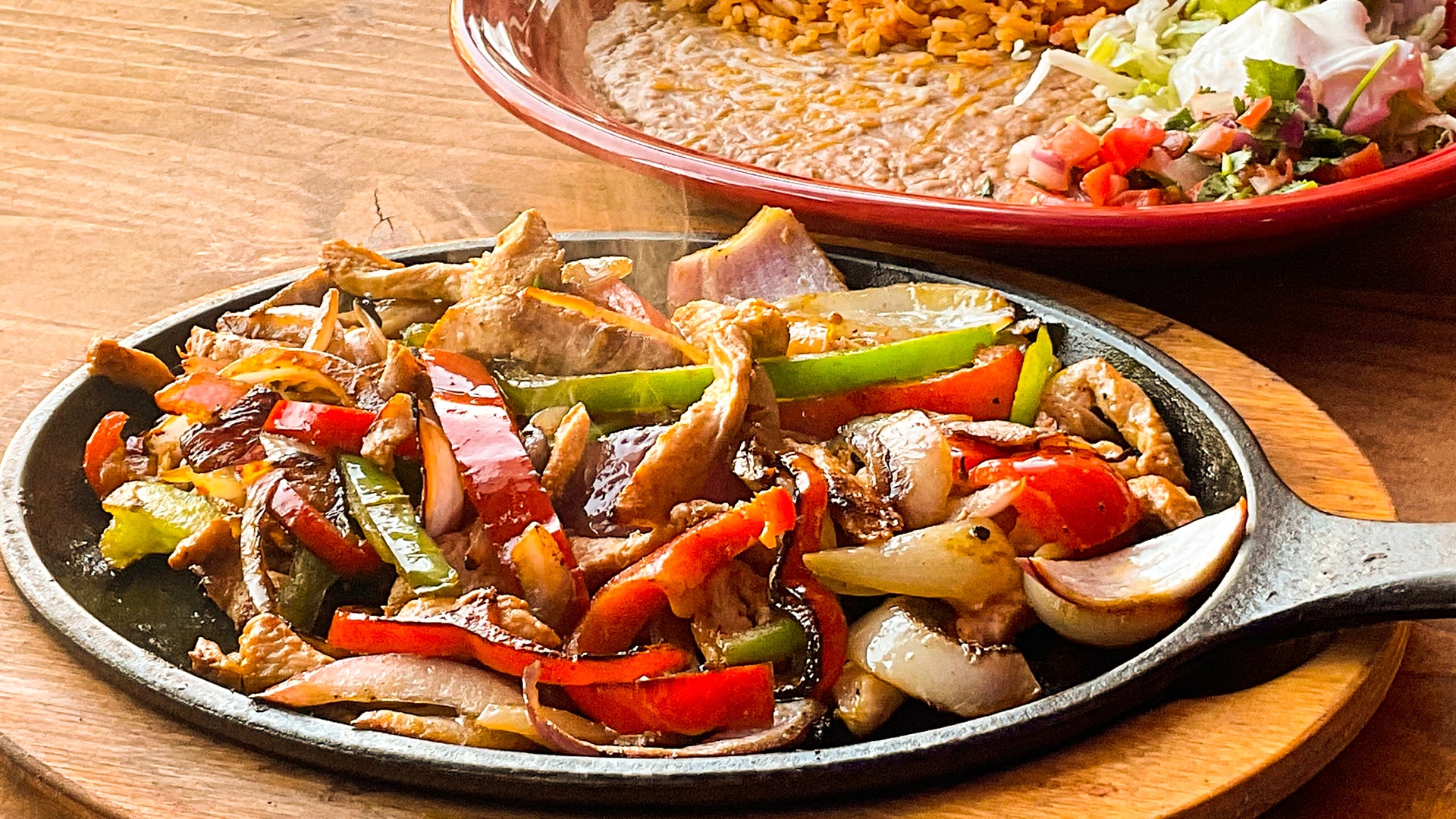Chicken fajitas steaming on a cast iron comal with peppers and onions, a plate of rice, beans and pico behind it